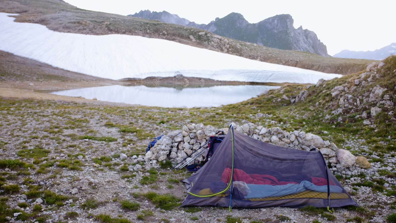 Backpacking tent pitched by a calm lake with snow-covered mountains